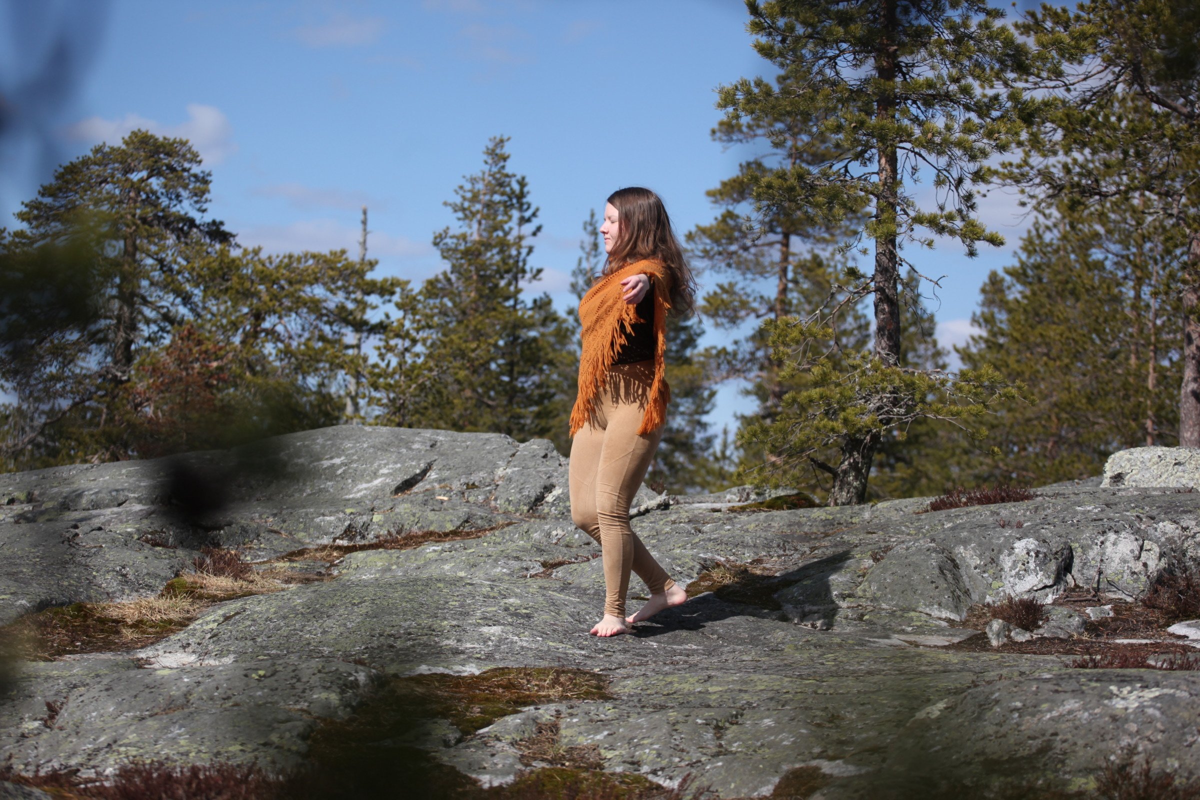 ihminen kalliolla oranssissa ponchossa ja vaaleanruskeissa housuissa, taustalla mäntyjä ja sininen taivas