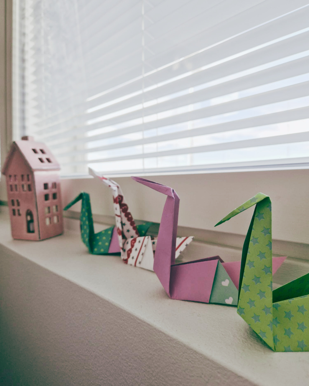 vihreitä ja vaaleanpunaisia taiteltuja paperikurkia ikkunalaudalla, taustalla vaaleanpunainen posliinitalo